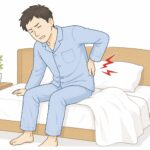 朝、ベッドから起き上がる際に腰を押さえて痛みを感じている男性のイラスト。寝起きの腰痛をイメージした画像。